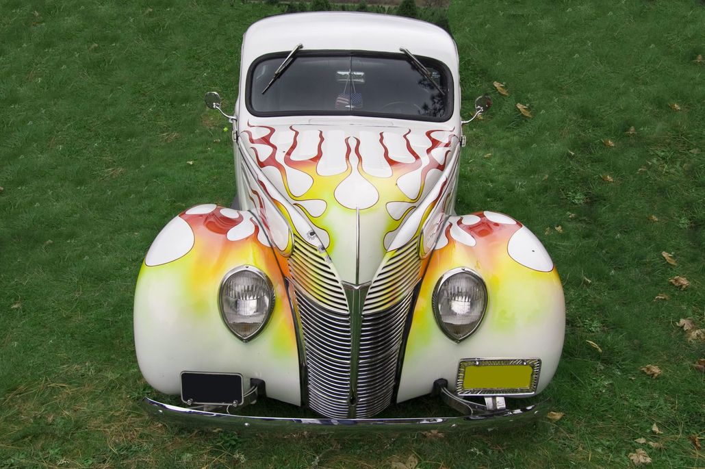 Custom-painted vintage car with white body, orange-yellow flames, and chrome grille on grass.