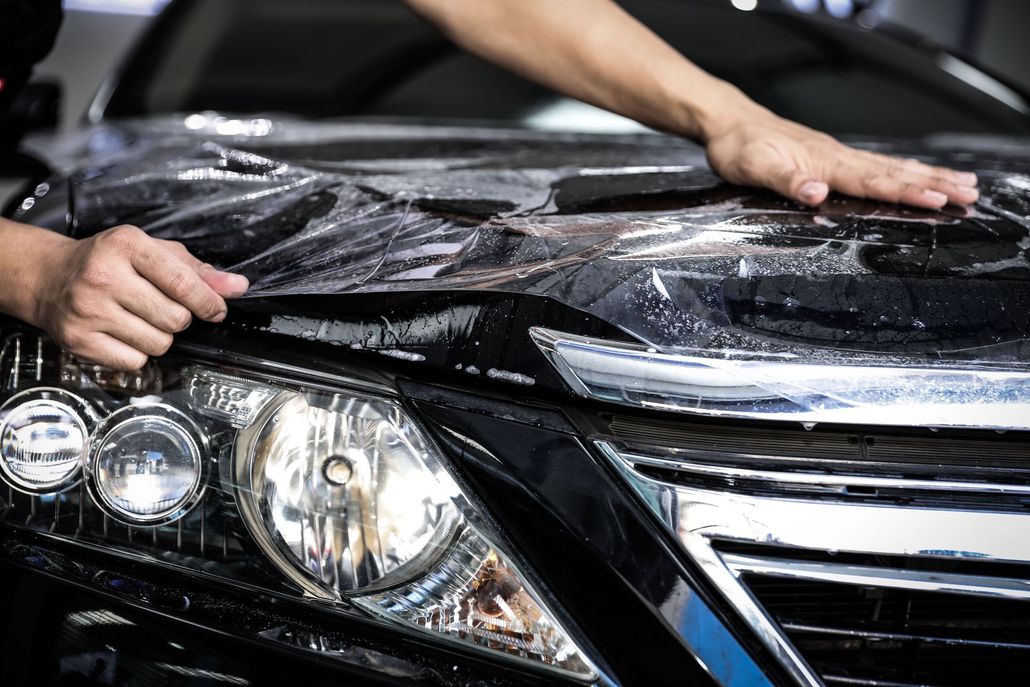 Hands washing a black car hood with soap suds, close-up on the front headlights and grille.