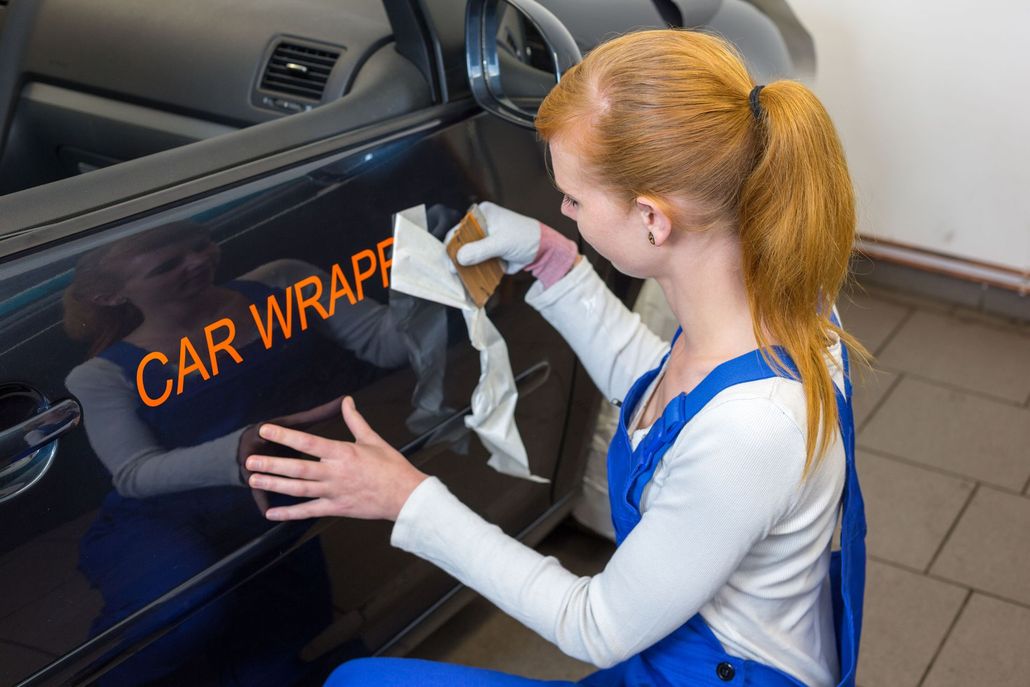 Woman applying a car wrap decal to a dark blue vehicle in a workshop.