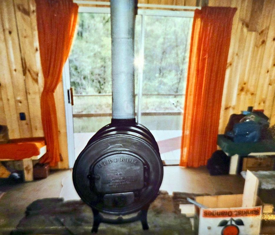 A wood-burning stove dominates a cabin interior with orange curtains and sliding glass door looking out.