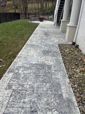 Gray patterned concrete walkway next to a house, with a grassy area and a rock bed.