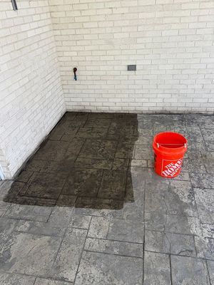 Stamped concrete patio, partially stained, with a Home Depot bucket next to a white brick wall.