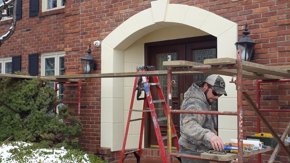 Man works on facade of red brick house, standing on scaffolding. Cream-colored trim surrounds the doorway.