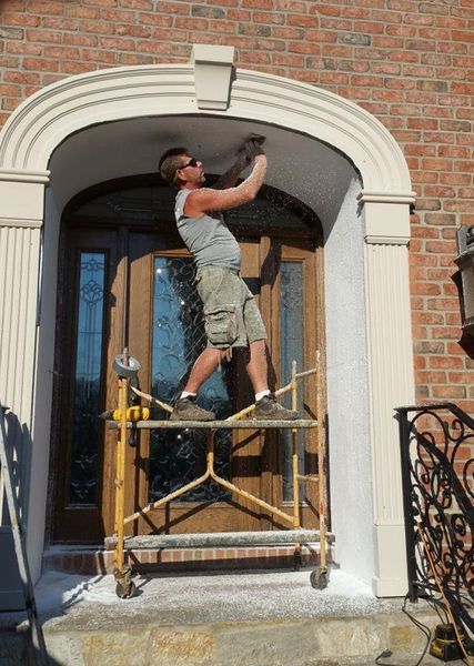Man on scaffold works on a building's arched doorway trim. He wears shorts, a tank top, and sunglasses.