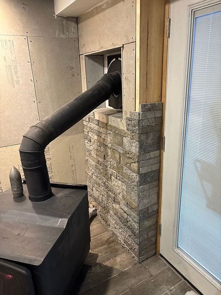 Wood-burning stove with black pipe, stone veneer wall, and doorway with blinds.