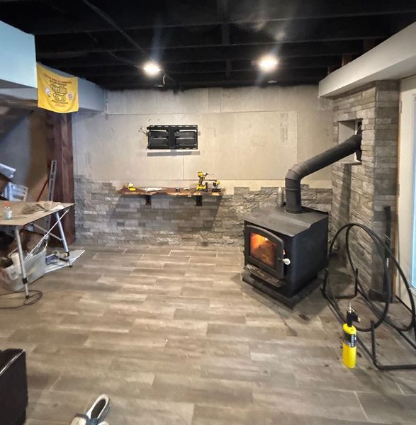 Basement with a wood-burning stove, tile floors, brick accent wall, and a partially finished ceiling.