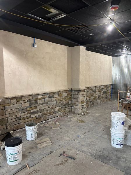 Interior construction site with stone wall and light beige stucco walls. Buckets and tools on concrete floor.