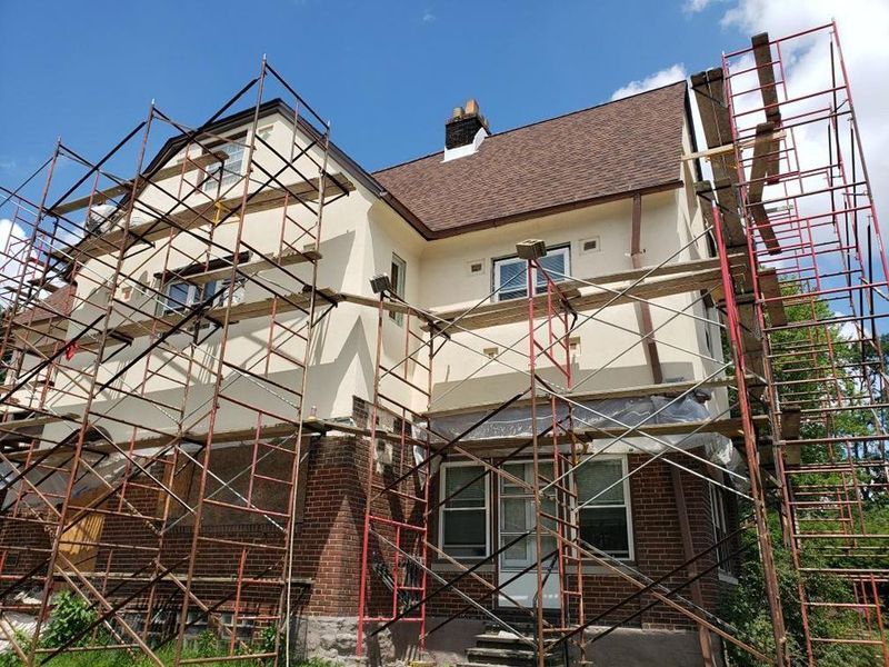 House with scaffolding, beige stucco, brown roof, brick facade, under construction, blue sky.