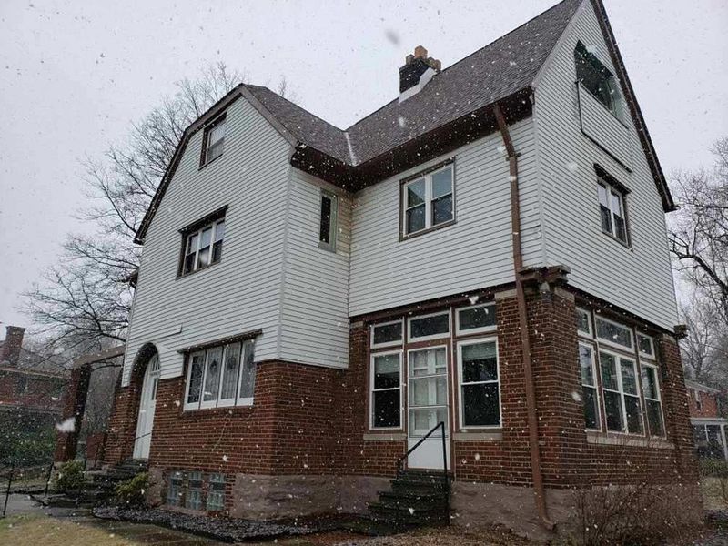 Two-story house with white siding, brick base, brown roof, and windows; it is snowing.