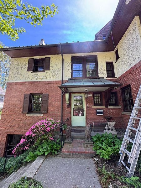 Two-story brick house with a green door, brown shutters, and pink flowers. A ladder leans against the side.