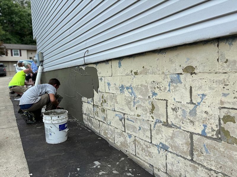 Two workers plastering a concrete foundation of a building; one kneels, applying cement.
