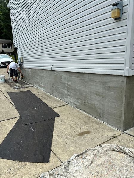 Person painting gray concrete foundation next to white vinyl siding of a house. Pavement, tarps, and a bucket are visible.