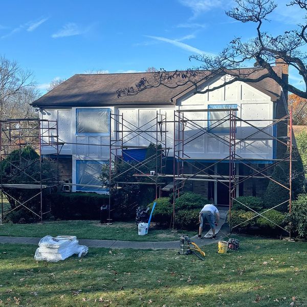 House exterior being painted, covered in protective plastic and scaffolding. A worker is visible.