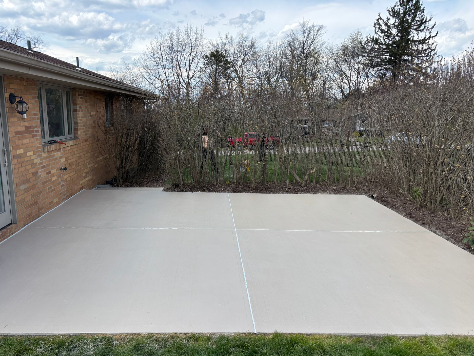 Concrete patio beside a brick house, with leafless shrubs and a fenced yard in the background