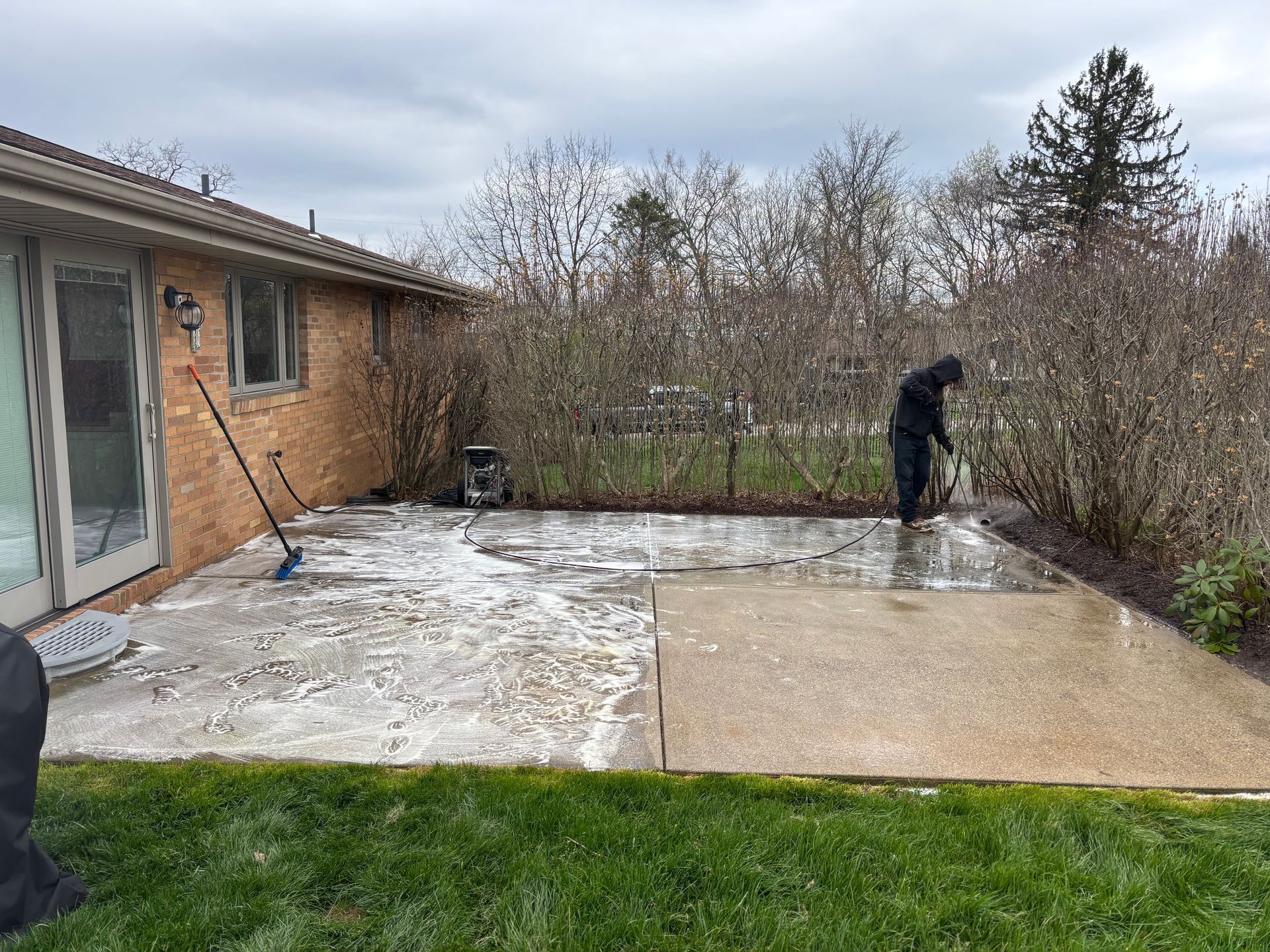 Person shoveling snow on a backyard patio beside a brick house and hedge