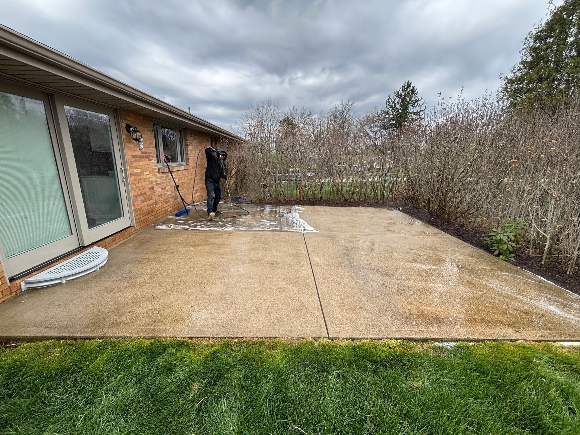 A person cleans a wet concrete patio behind a brick house under a cloudy sky. 