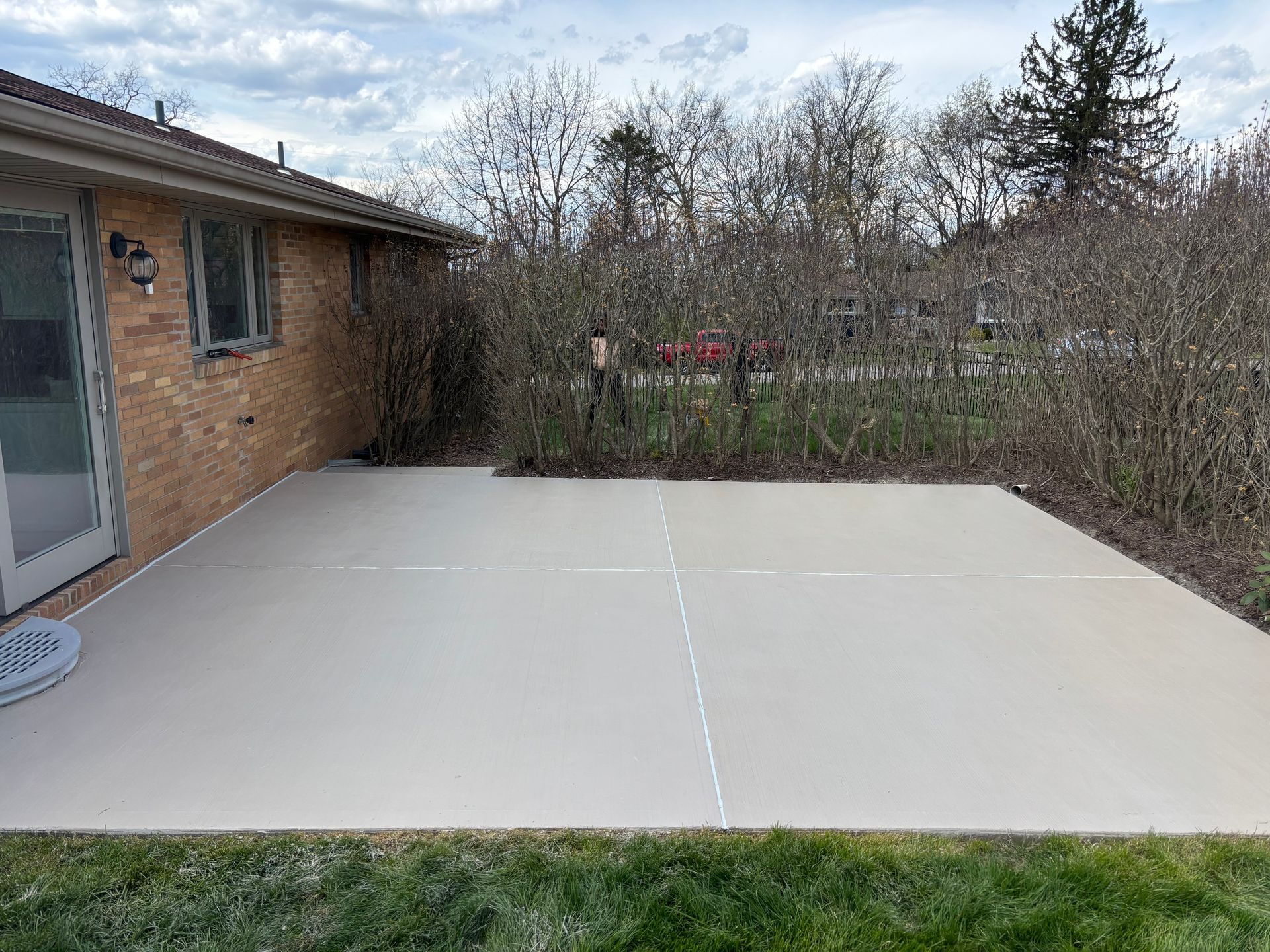 A newly poured, light gray concrete patio sits adjacent to a tan brick house, bordered by bare shrubs and a grassy lawn. 