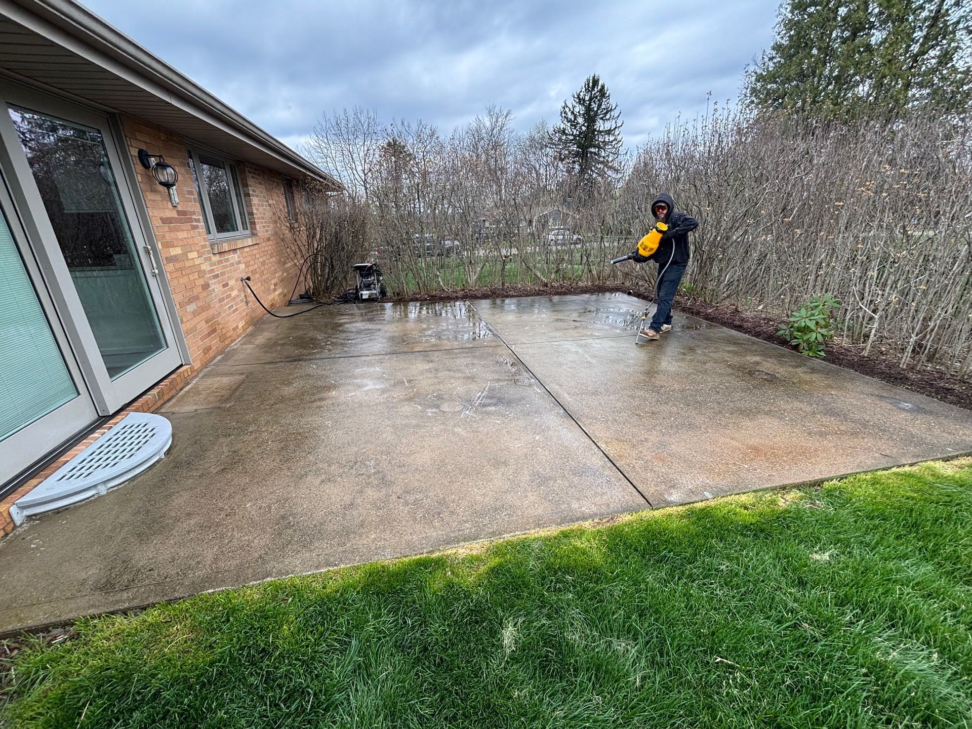 A person in dark clothing uses a yellow power washer to clean a concrete patio next to a brick house. 