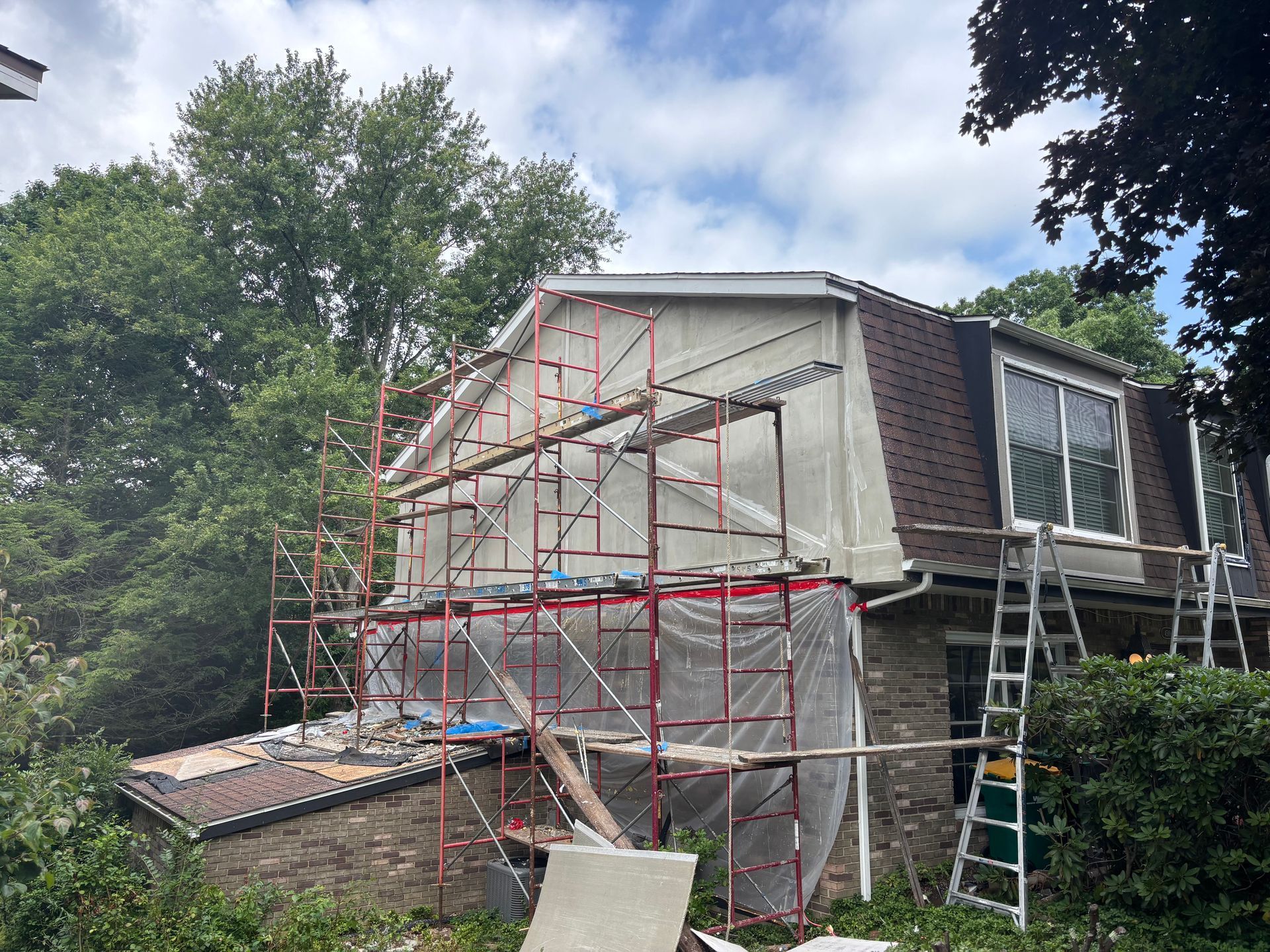 House under renovation with red scaffolding, white tarp, and ladder beside trees
