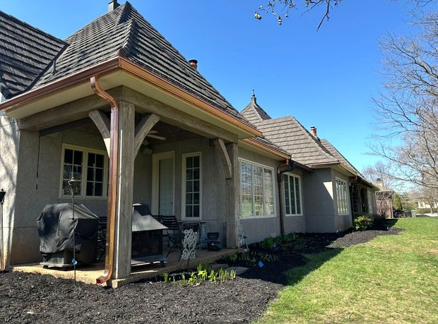 Covered patio on a stucco house with two grills, dark roof tiles, and a grassy yard under a clear blue sky