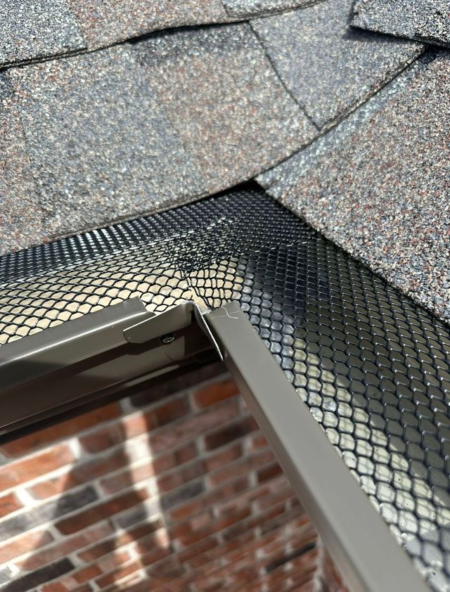 Close-up of a shingled roof edge with black metal flashing above a brick wall.