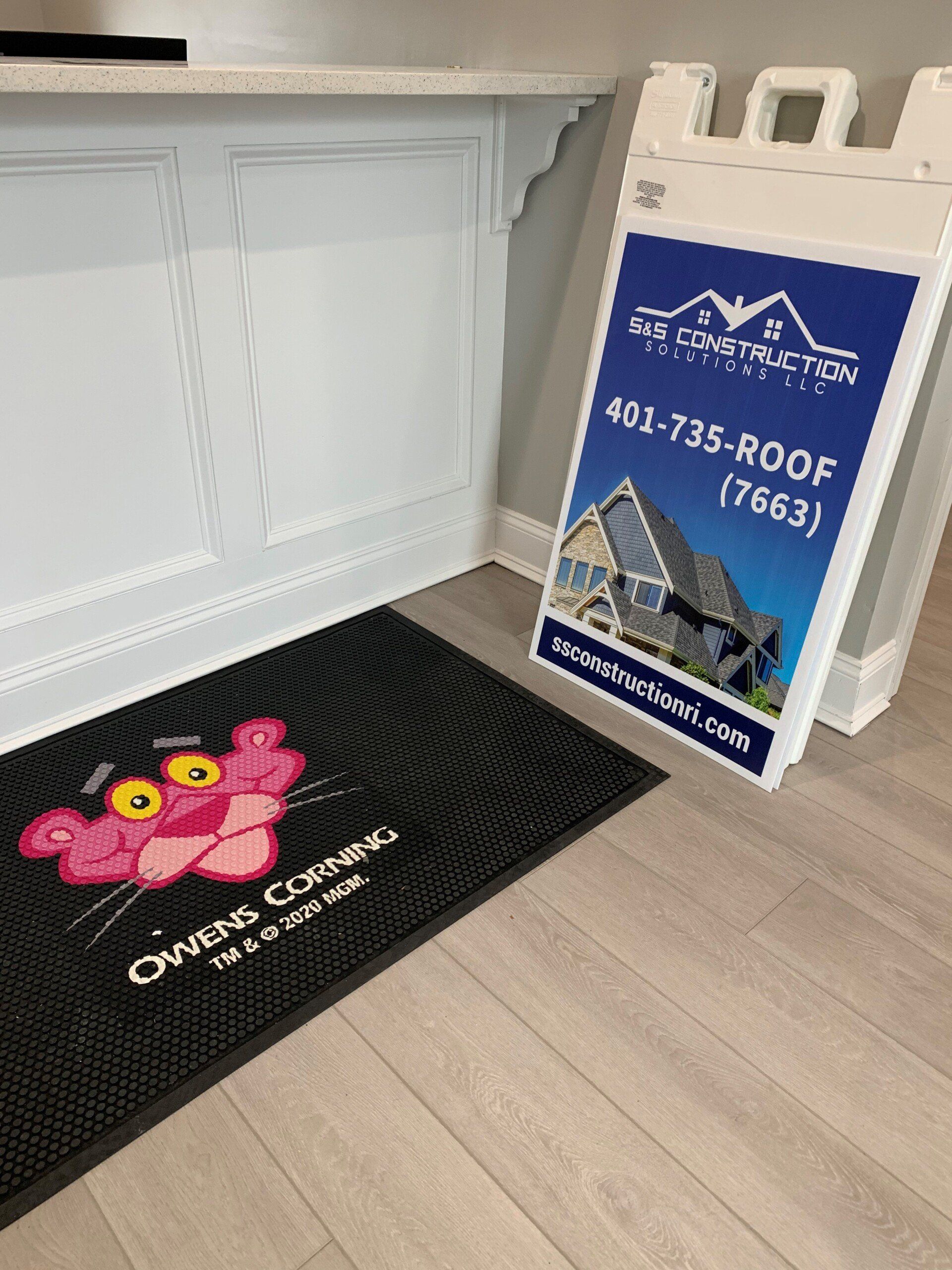 A pink panther doormat and a blue and white sign for roofing services stand in a corner. The setting is indoors.