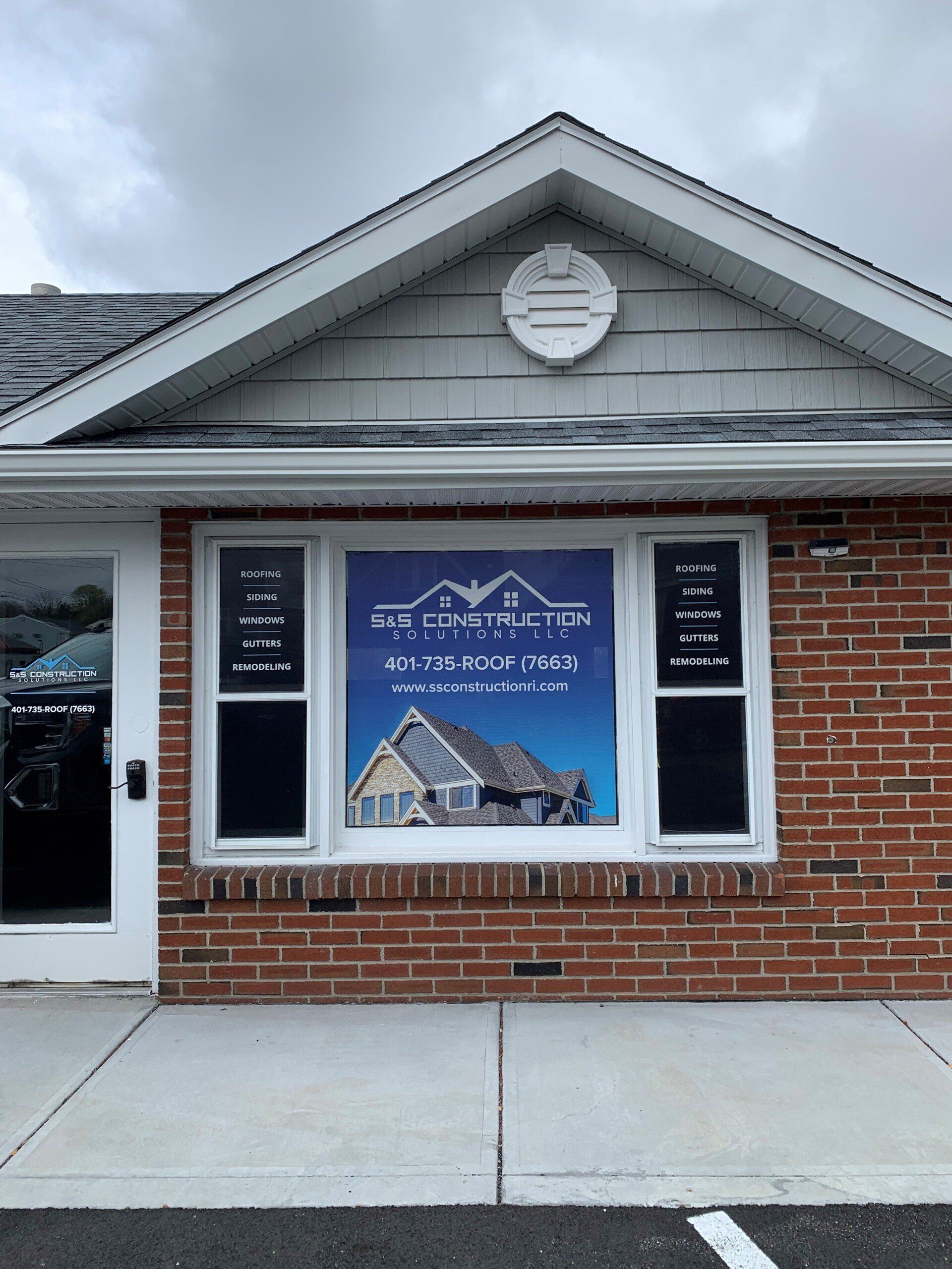 Exterior of a brick building with a large window displaying a real estate sign. The sign features a photo of a house and text.