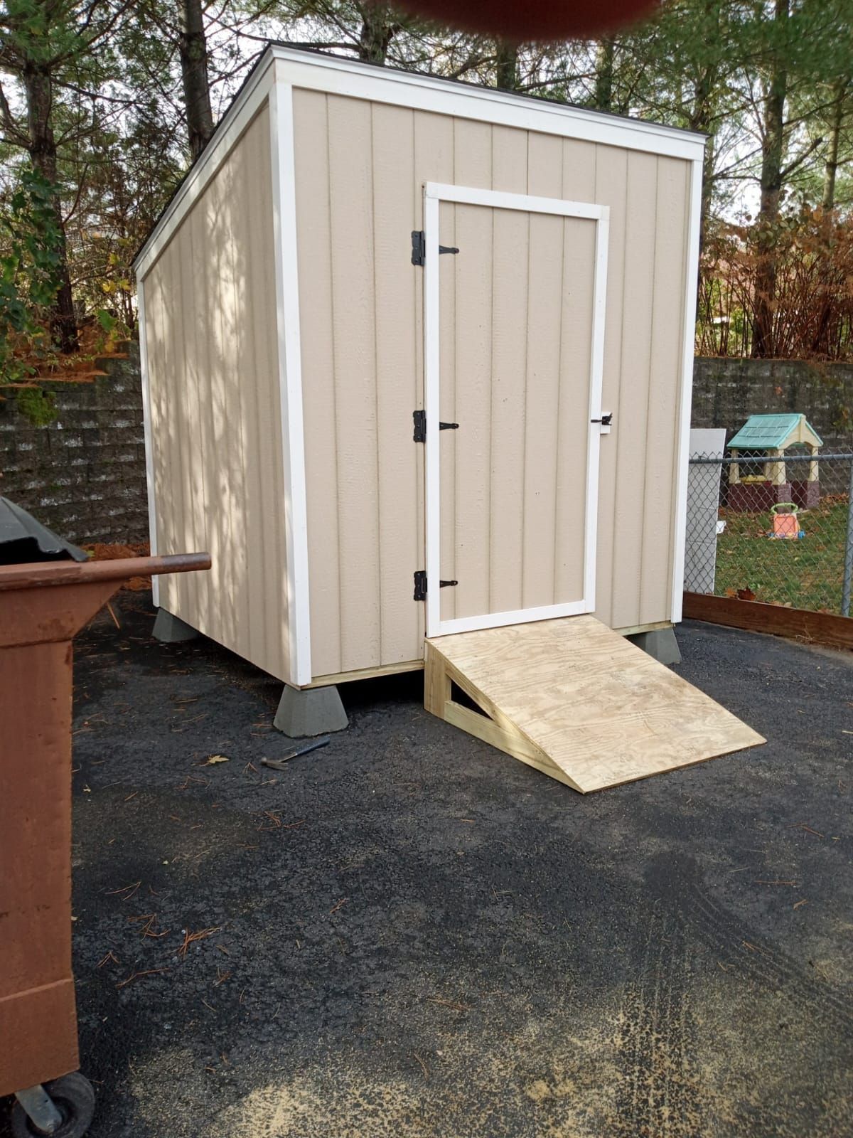 A beige shed with a wooden ramp in a backyard. The shed has a white-framed door and is raised on concrete blocks.