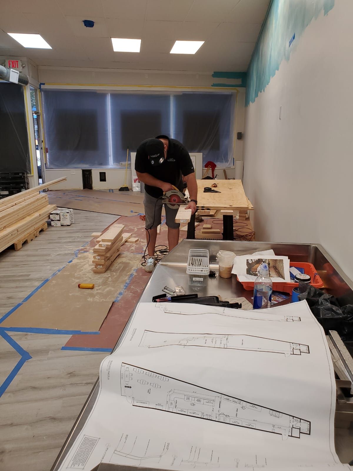 A person uses a saw to cut wood inside a room under renovation. Blue tape outlines areas on the floor; blueprints and tools are nearby.