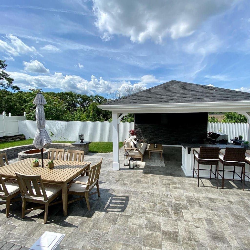 Outdoor patio with dining table, bar, and seating under a gazebo. White fence and blue sky with clouds.