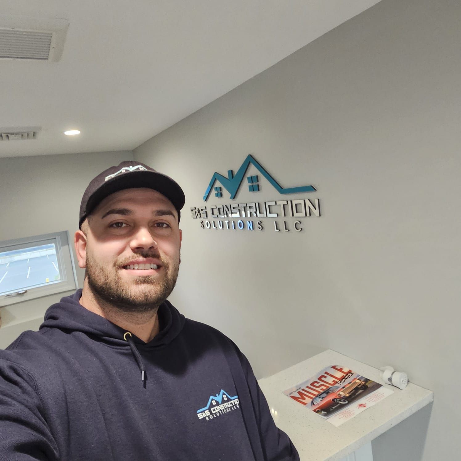 Man in a baseball cap and sweatshirt smiles in front of a company logo on a wall. He is standing inside a light-colored room.