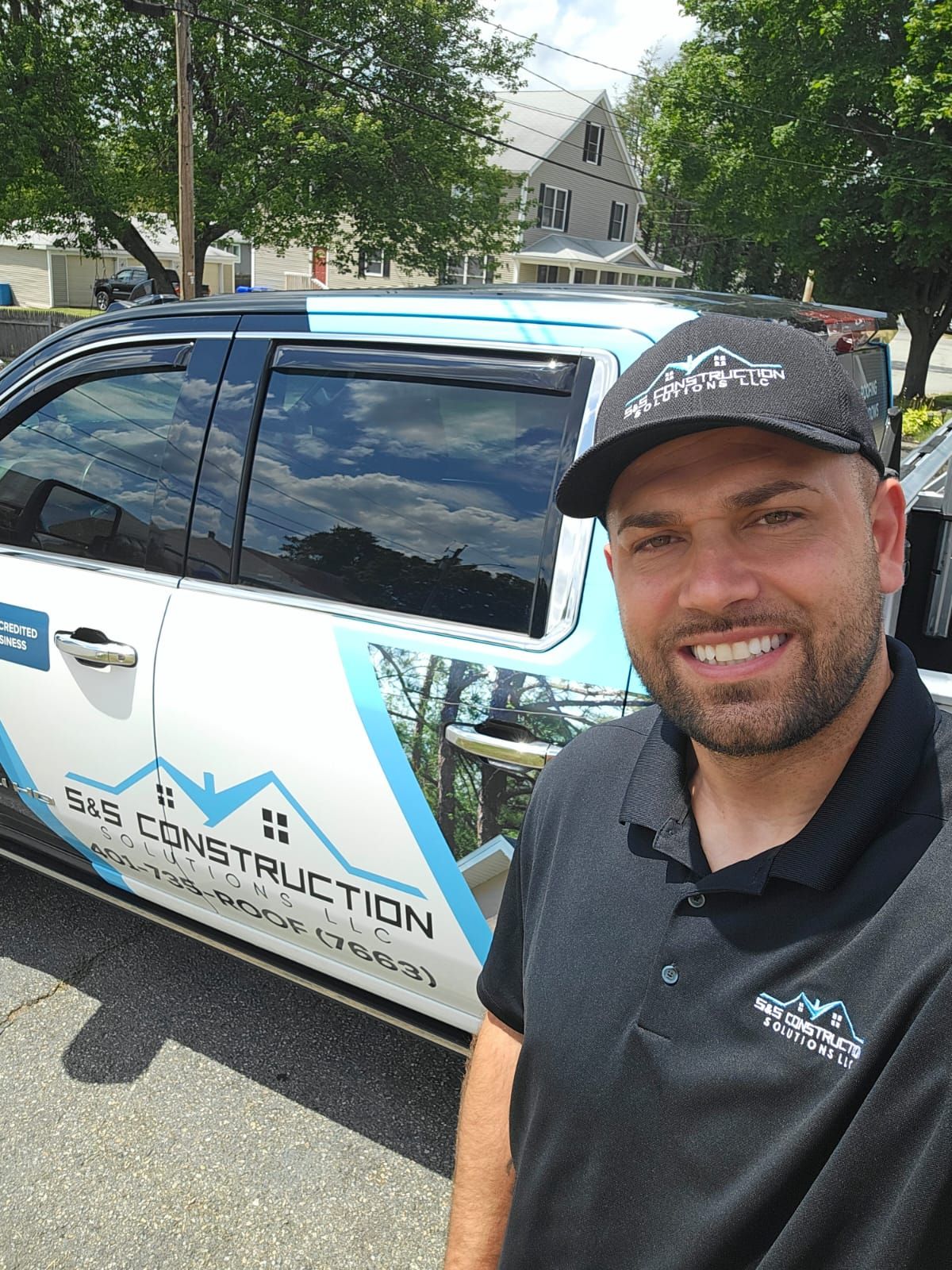 Man in black shirt and hat with BVE Construction logo smiling next to a branded vehicle on a sunny day.