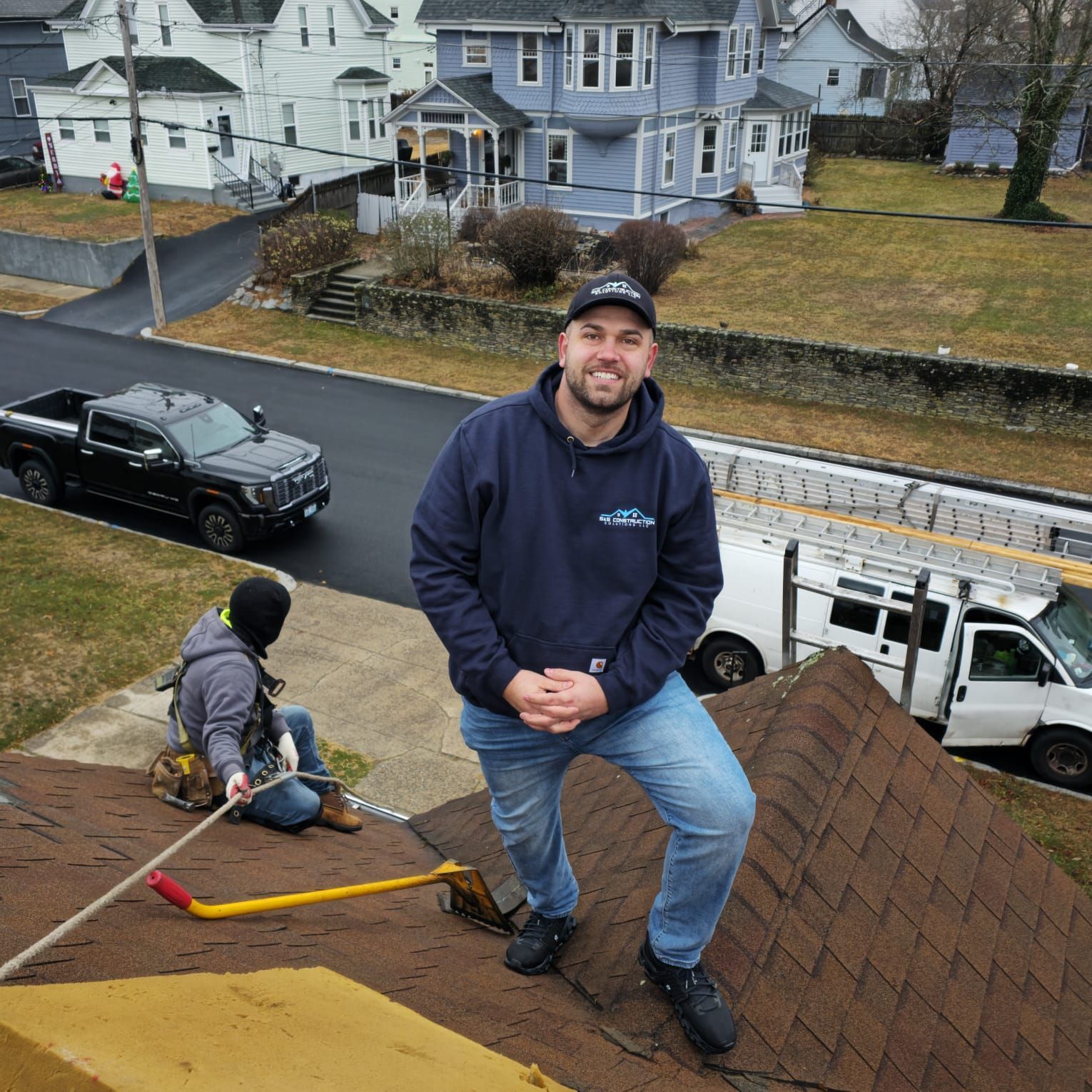 Man on a roof smiles at the camera, another worker is secured with a rope. Houses and a black truck are in the background.