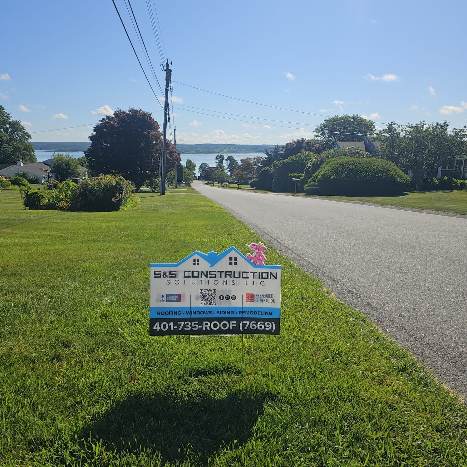 A sign for S & S Construction Solutions, LLC in a grassy yard next to a road leading to a body of water. Bright, sunny day.