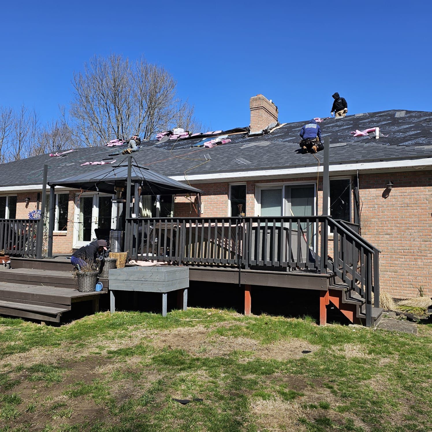 Roofers working on a brick house roof, removing old shingles. Clear blue sky in the background, sunny day.