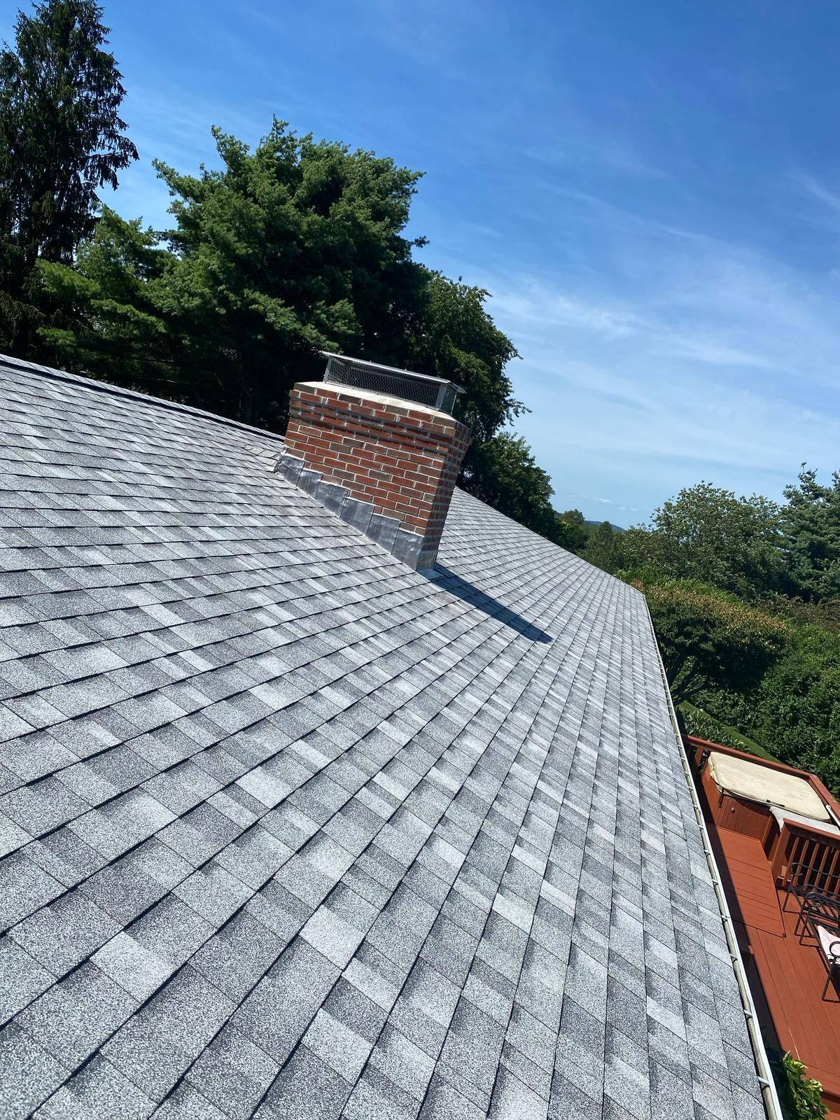 Gray shingled roof with brick chimney on a sunny day. Trees and a deck are visible in the background.