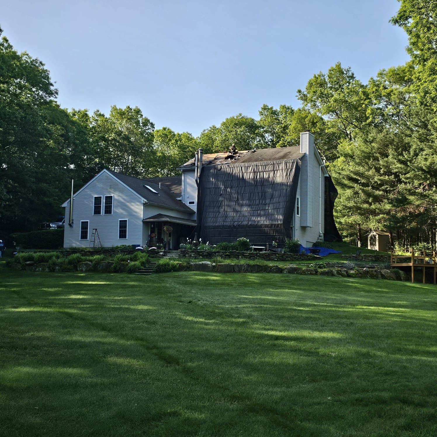 A two-story house with a damaged roof, surrounded by trees and a green lawn under a blue sky.