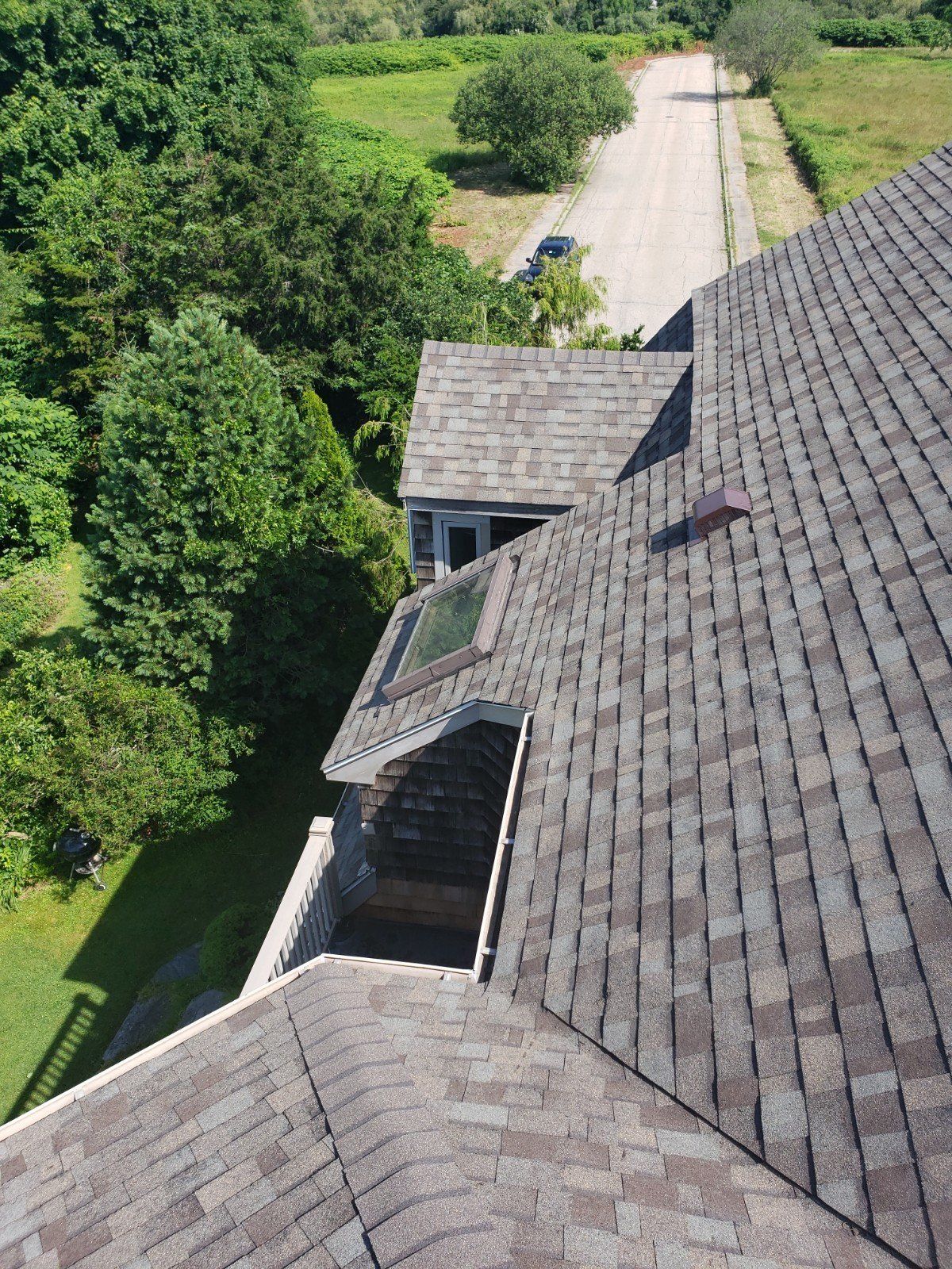 Aerial view of a house roof with shingles, a skylight, and a road in the background surrounded by green trees.