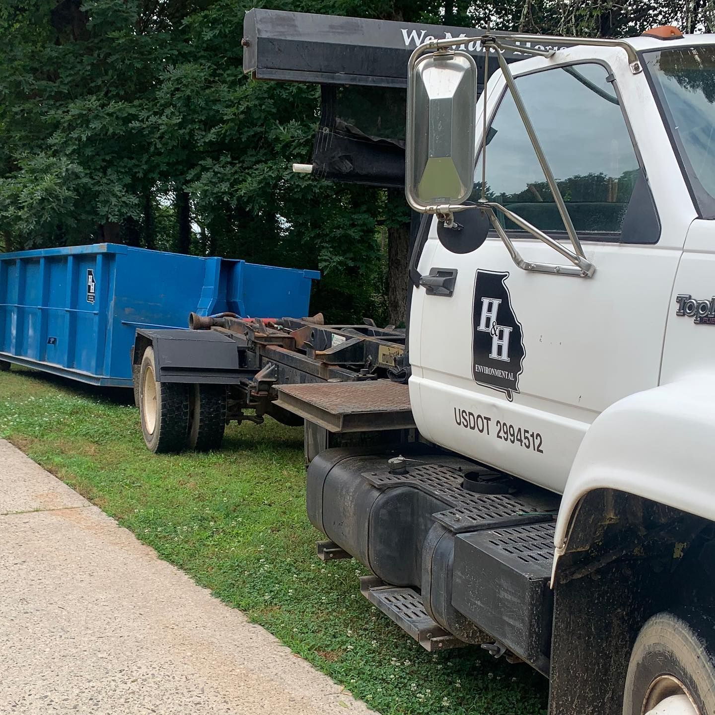 White tow truck with flatbed parked beside a blue dumpster on a grassy roadside near trees.