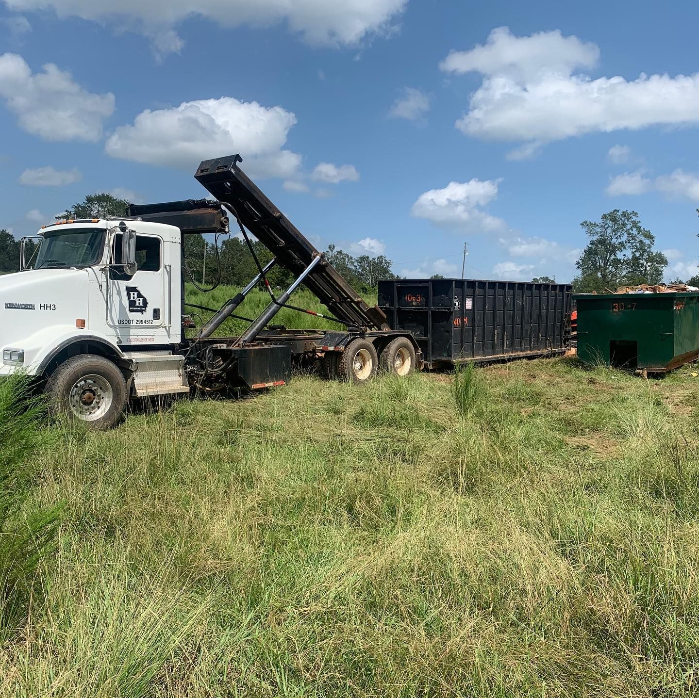 White dump truck loading debris into a black trailer in a grassy field under a blue sky