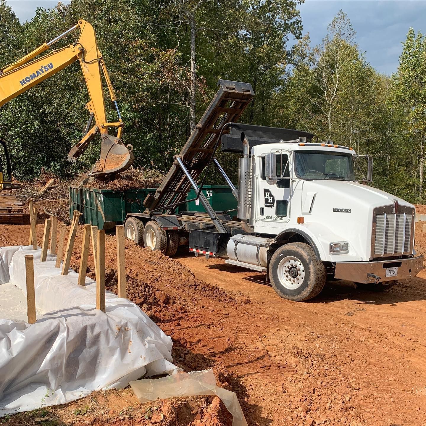 Excavator loading dirt into a dump truck at a construction site with red clay soil.