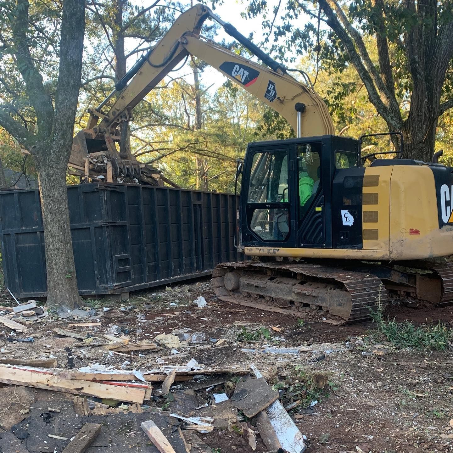 Yellow excavator loading debris into a black dumpster in a wooded lot