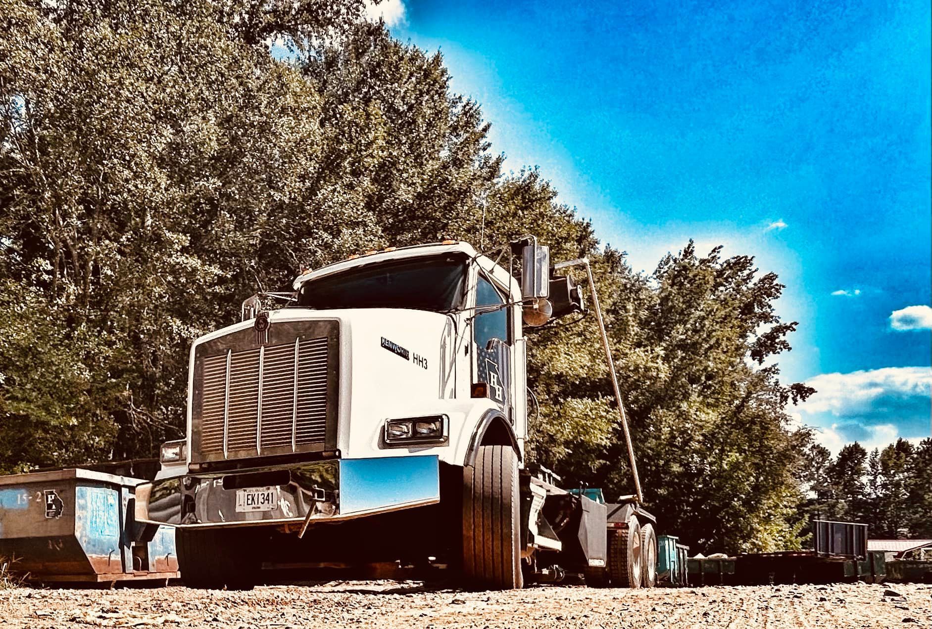 White dump truck on a dirt road under a bright blue sky, with trees in the background
