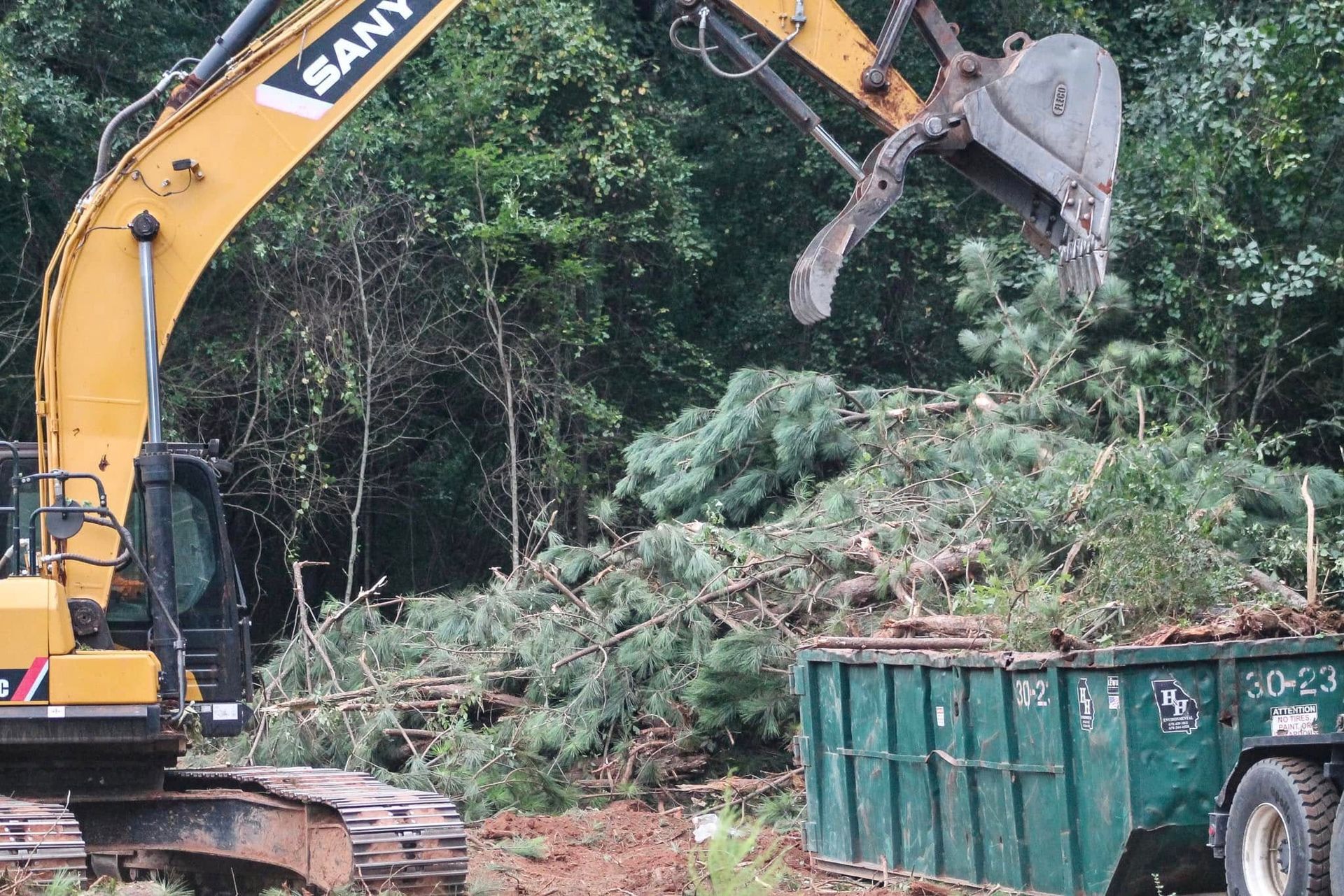 Yellow excavator loading green brush into a dumpster in a wooded area