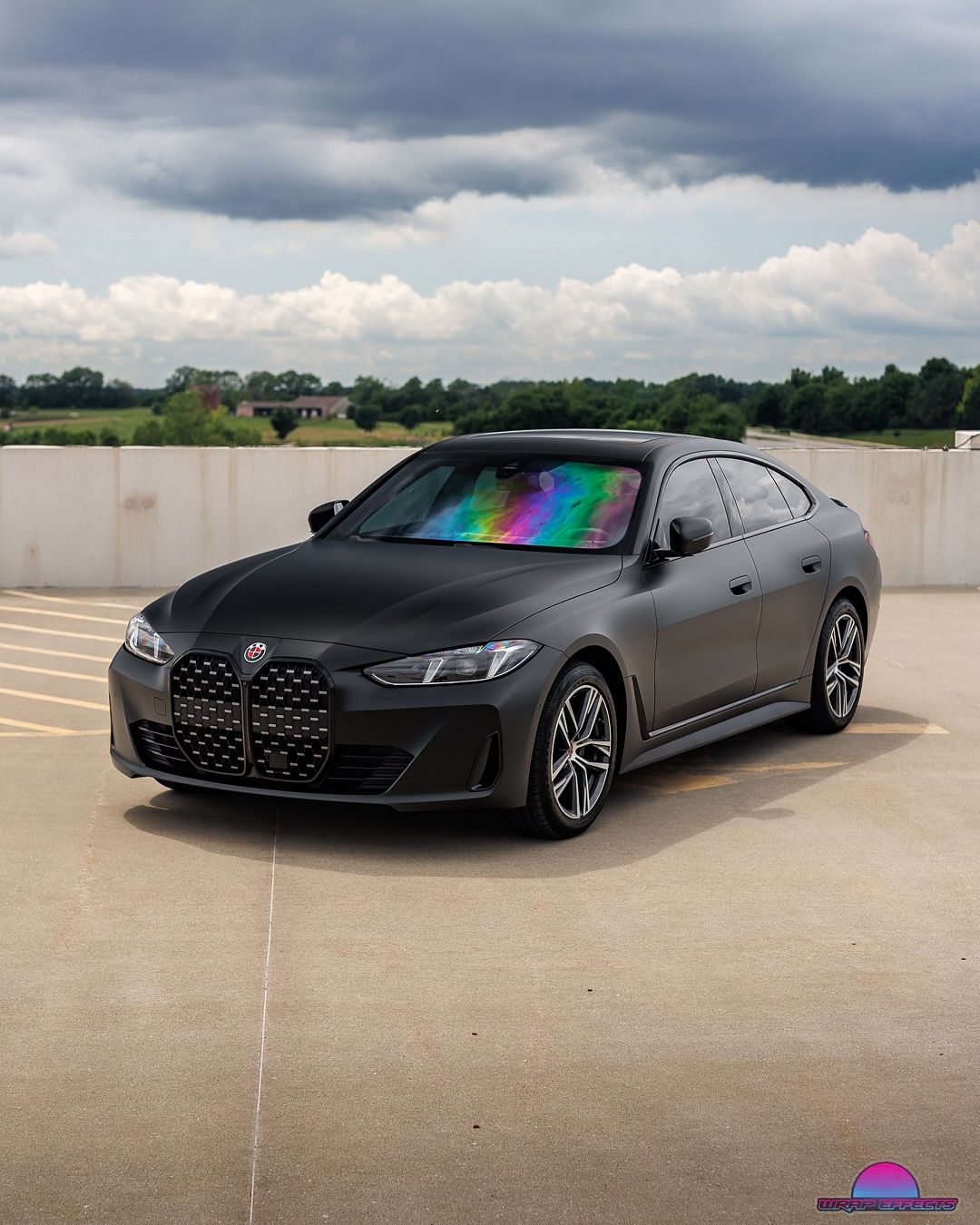 Dark gray BMW i4 parked on a rooftop, with rainbow-colored sunshade, against a cloudy sky.