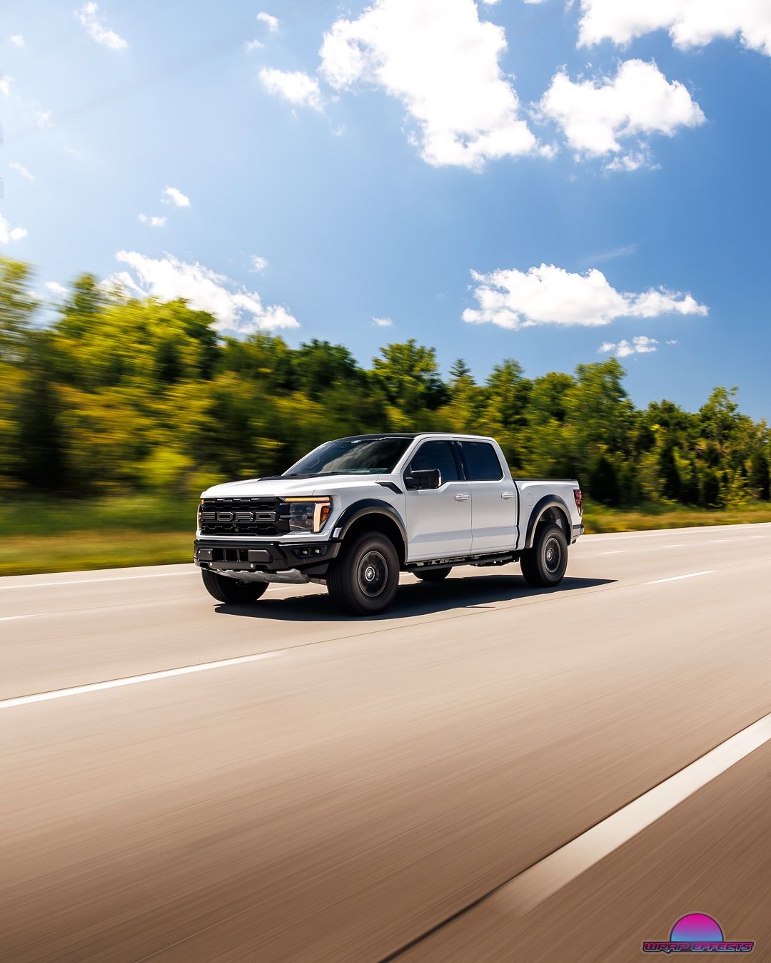 White Ford Raptor truck driving on a highway, trees in background, blue sky with clouds.