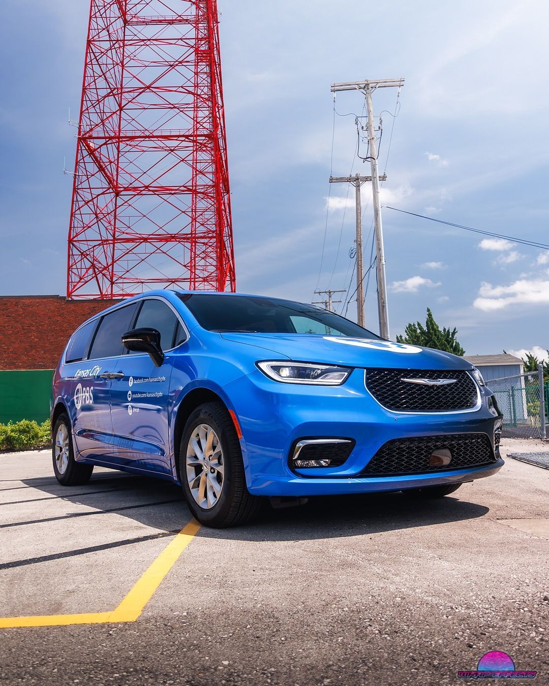 Blue Chrysler Pacifica minivan parked in lot, with red tower and blue sky in background.