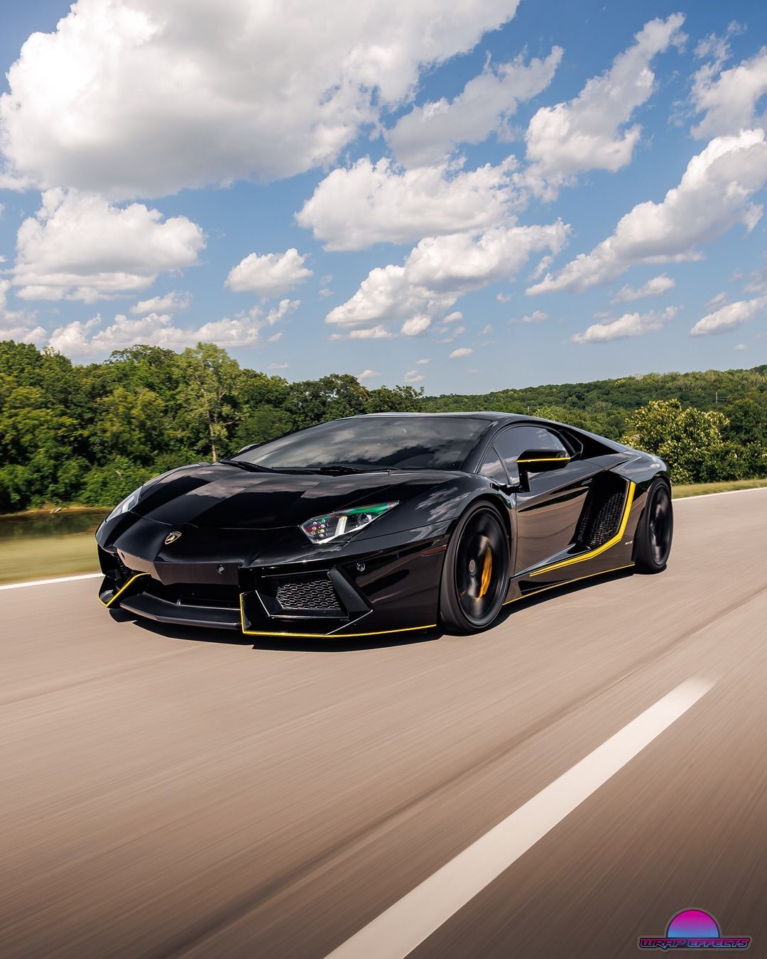 Black Lamborghini Aventador on a paved road, yellow accents, blue sky, trees in background.
