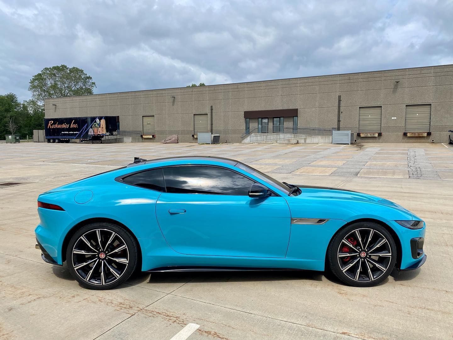 A bright blue Jaguar F-Type sports car parked on a concrete lot in front of a warehouse.