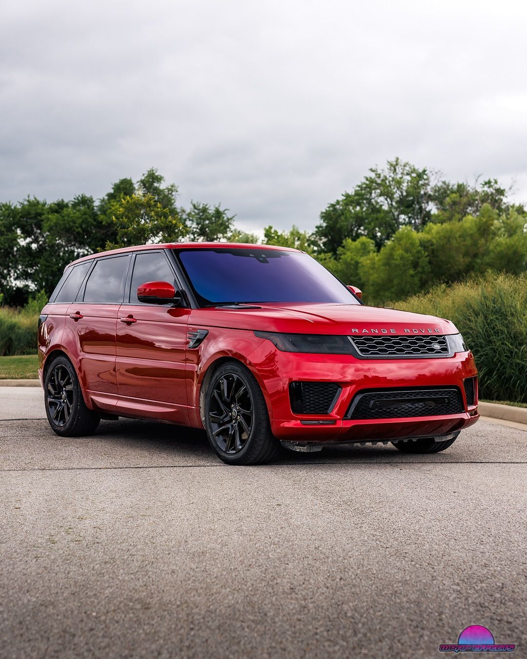 Red Range Rover Sport on a paved surface with black wheels, against a background of green trees and an overcast sky.